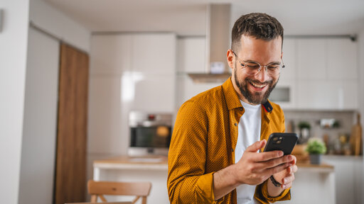 Mann mit Brille steht in der Küche und schaut fröhlich auf das Handy